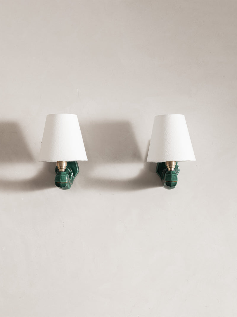 Two matching wall sconces mounted side by side on a light beige wall. Each sconce has a white tapered lampshade, a green sculptural wall mount, and a brass socket.