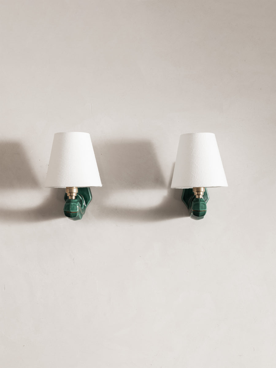 Two matching wall sconces mounted side by side on a light beige wall. Each sconce has a white tapered lampshade, a green sculptural wall mount, and a brass socket.